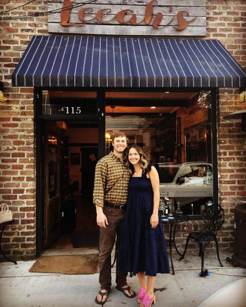 A couple stands outside a restaurant named "Leah's," smiling in front of a brick wall and a striped awning.