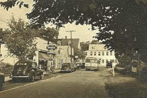 A vintage street scene featuring old cars, a bus, and small shops under trees, evoking a nostalgic atmosphere.