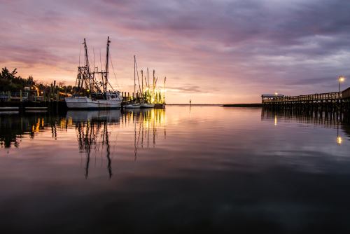 A serene harbor at sunset, featuring fishing boats and a calm water reflection under a colorful sky.