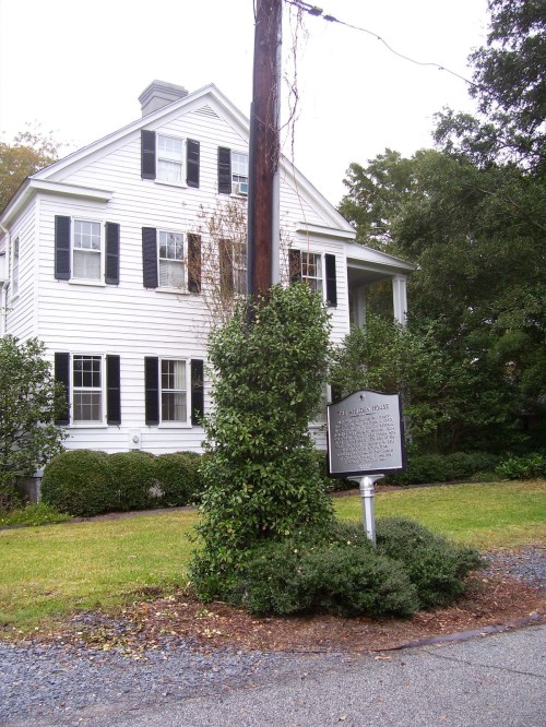 A white two-story house with black shutters, surrounded by greenery and a historical marker in front.