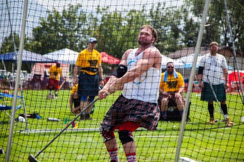 A man in a kilt swings a heavy hammer in a sports competition, with spectators in the background under colorful tents.