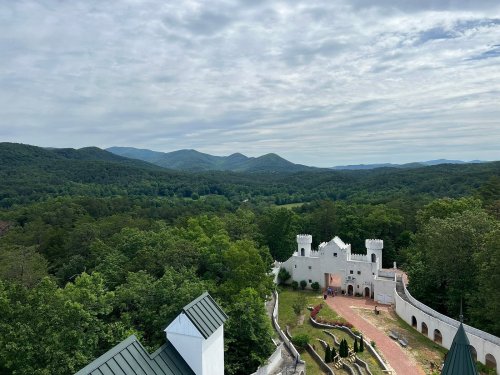 A scenic view of a white castle surrounded by lush greenery and mountains under a cloudy sky.
