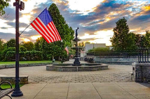 A park scene featuring a fountain, American flags, and a colorful sunset sky.