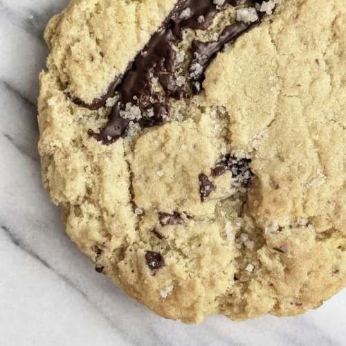 Close-up of a chocolate chip cookie with a cracked surface and coarse sugar on top, resting on a marble countertop.