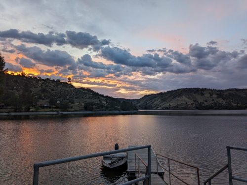 Sunset over a calm lake, with clouds reflecting in the water and hills in the background. A small boat is docked nearby.