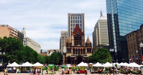 A city square with tents and vendors, featuring a historic church and modern skyscrapers in the background.