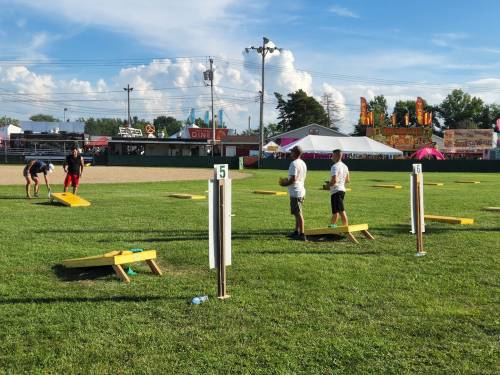People playing cornhole on a grassy field with a fair in the background and blue skies above.