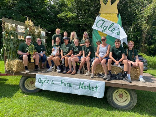 A group of people in green shirts poses on a trailer decorated with corn and a sign for Agle's Farm Market.