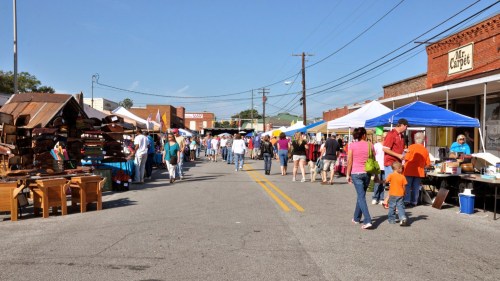 A bustling street market with vendors under colorful tents, people browsing, and sunny skies overhead.