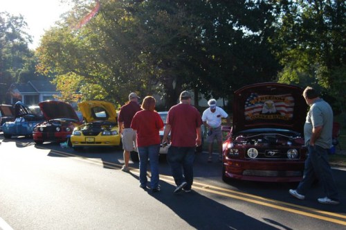 A group of people walking by classic cars with hoods open, showcasing their engines on a sunny day.