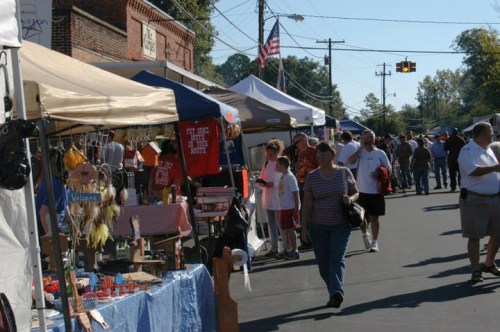 A bustling street market with vendors under tents, people browsing, and an American flag in the background.