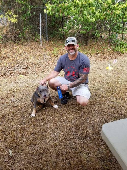 A smiling man in a gray shirt kneels beside a dog in a grassy yard, surrounded by trees and a fence.