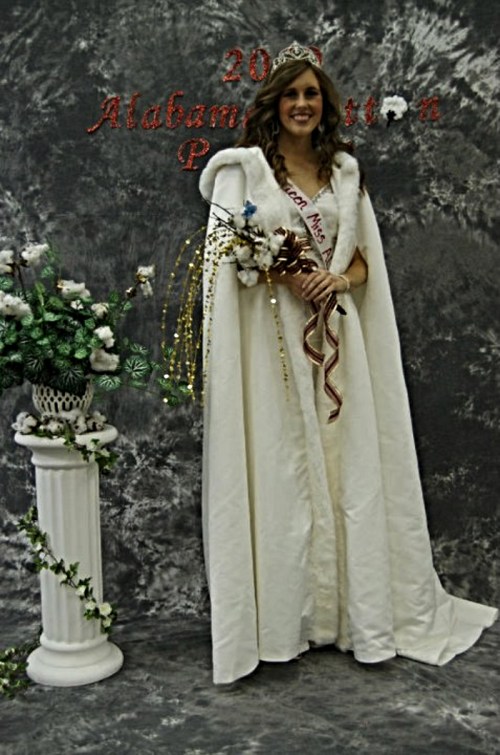 A young woman in a white gown and crown stands beside a decorated column, holding a bouquet and wearing a sash.
