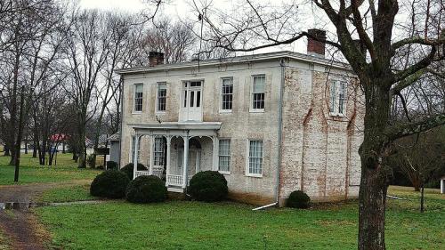 A historic two-story white brick house with a porch, surrounded by trees and a grassy yard on a cloudy day.