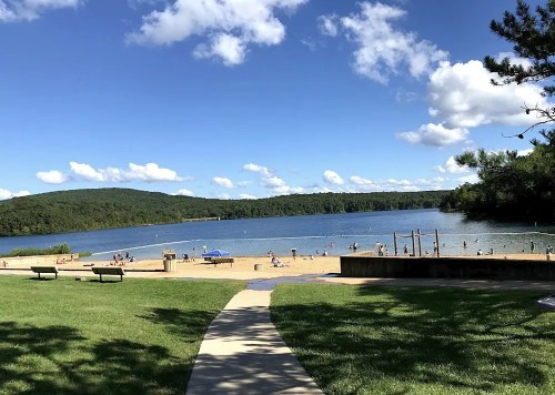 A scenic view of a sandy beach by a lake, surrounded by green hills and dotted with people enjoying the water on a sunny day.