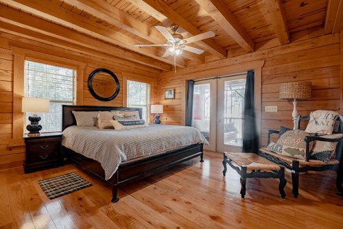 Cozy wooden bedroom with a bed, two lamps, a chair, and large windows, featuring rustic decor and warm lighting.