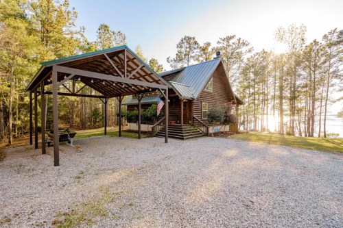 A rustic cabin with a metal roof, surrounded by trees and a gravel driveway, near a lake at sunset.