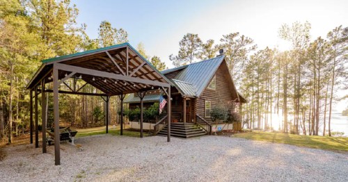 A rustic cabin with a metal roof, surrounded by trees and a lake, featuring a covered porch and gravel driveway.