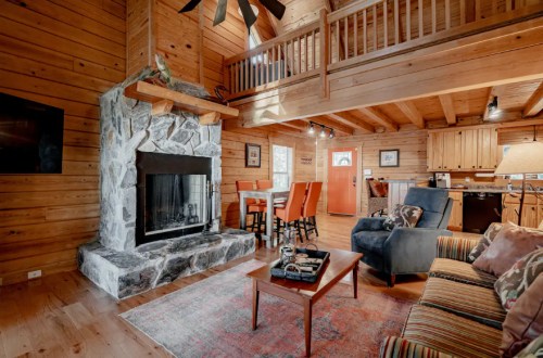 Cozy wooden cabin interior with a stone fireplace, seating area, and dining space, featuring warm lighting and rustic decor.