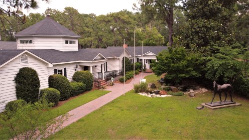 Aerial view of a white house with a circular tower, surrounded by greenery and a horse statue in the garden.
