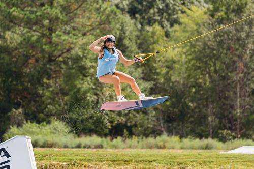 A woman in a helmet jumps on a wakeboard, holding a rope, with greenery in the background.