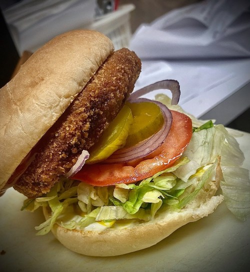 A close-up of a sandwich with a crispy patty, lettuce, tomato, pickles, and onions on a soft bun.