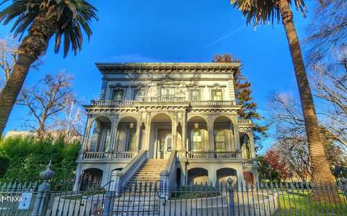 A grand, historic mansion with a gray facade, large columns, and palm trees in front, set against a clear blue sky.