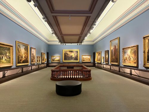 A spacious art gallery featuring large framed paintings on blue walls and a central seating area.