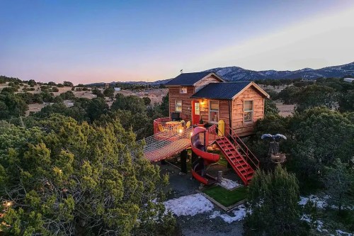 A wooden treehouse with a slide and stairs, surrounded by trees at sunset, showcasing a playful and cozy design.