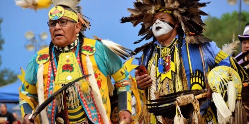Two Indigenous men in traditional attire, adorned with feathers and vibrant colors, participate in a cultural ceremony.