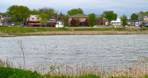 View across the lake toward the town of Cumberland, Wisconsin, featuring green trees and storefronts.