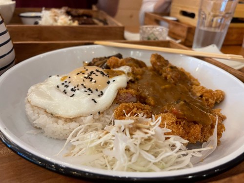 A plate of rice topped with a fried egg, crispy chicken, curry sauce, and shredded cabbage, served with chopsticks.