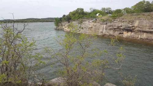 A serene river scene with rocky cliffs, green trees, and a camping area in the background under a cloudy sky.
