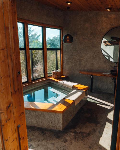 A modern bathroom featuring a sunlit soaking tub, wooden accents, and large windows with a natural view.