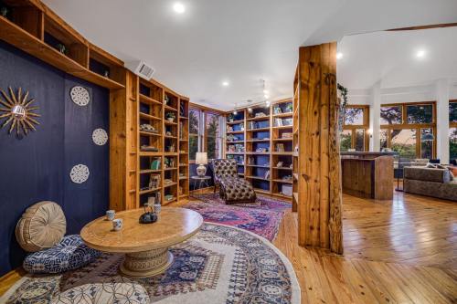 Cozy living space with wooden shelves, a patterned rug, and a round table, featuring large windows and warm lighting.