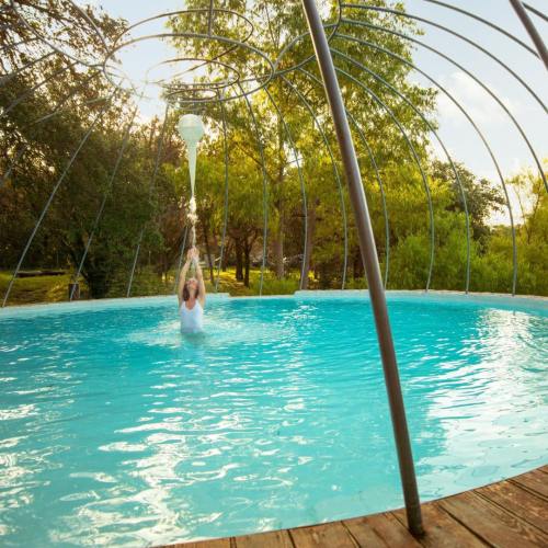 A person swings from a structure above a clear blue pool surrounded by greenery under a sunny sky.