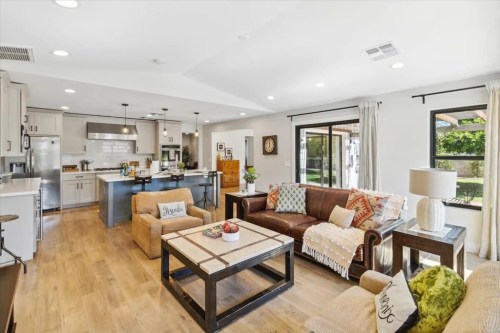 A modern living room with a brown leather sofa, coffee table, and open kitchen, featuring bright natural light and greenery outside.