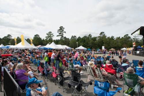 A large crowd gathers at an outdoor event with tents and a stage, seated in folding chairs under a cloudy sky.