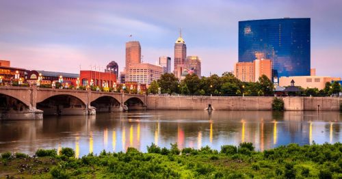 Evening views of the skyline of Indianapolis, reflecting on the river, with lush greenery in the foreground.