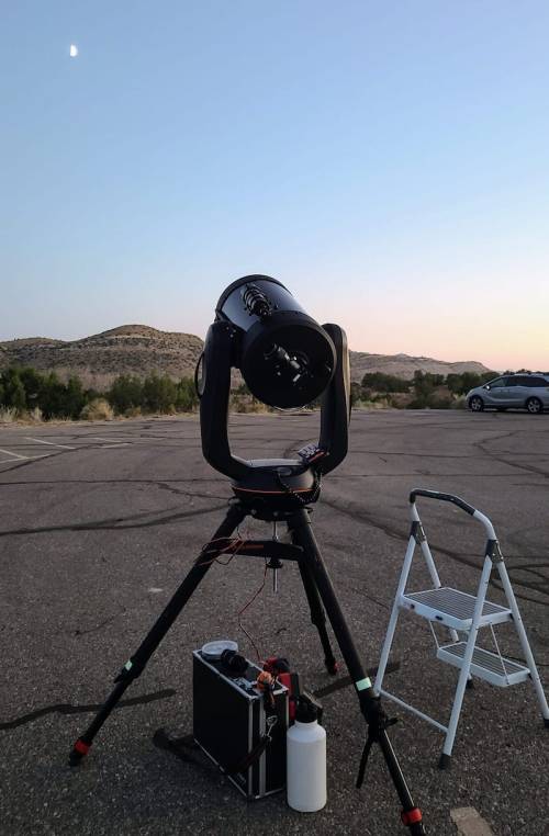 A telescope set up on a tripod in a parking area, with mountains in the background and a crescent moon in the sky.