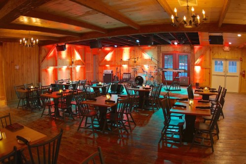 Cozy restaurant interior with wooden decor, tables set for dining, and a stage with musical instruments in warm lighting.