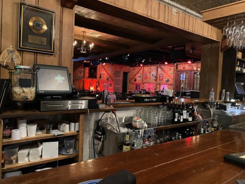 A rustic bar with wooden decor, shelves of drinks, and a stage in the background illuminated by red lights.