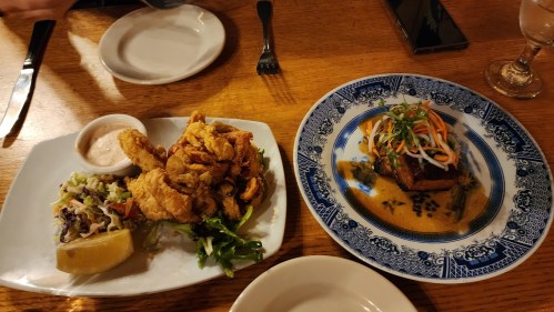A plate of fried chicken with coleslaw and a lemon wedge, alongside a dish of fish in sauce topped with vegetables.