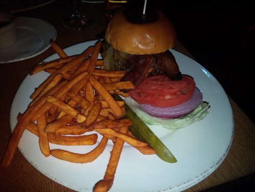 A juicy burger topped with bacon, served with lettuce, tomato, onion, pickles, and a side of crispy sweet potato fries.