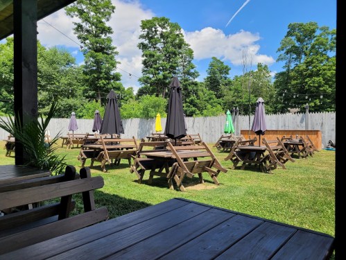 Outdoor seating area with wooden tables and umbrellas, surrounded by green grass and trees under a blue sky.
