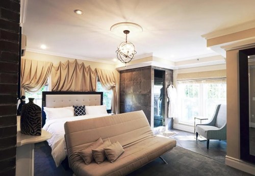 A stylish bedroom featuring a bed, elegant drapes, a cozy chair, and a modern bathroom area with a glass shower.
