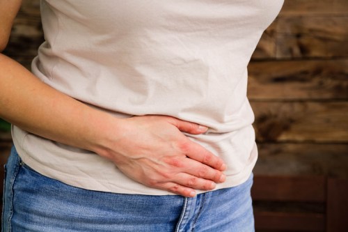 A person in a light shirt and jeans holds their stomach, indicating discomfort, against a wooden background.