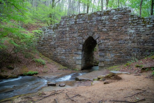 A stone arch bridge over a flowing stream, surrounded by lush green trees and a forested landscape.