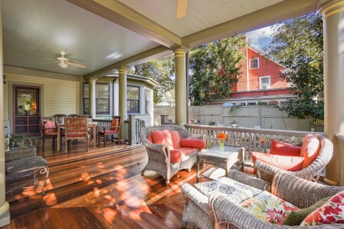 A cozy porch with wicker furniture, red cushions, and a dining area, surrounded by greenery and a charming house.