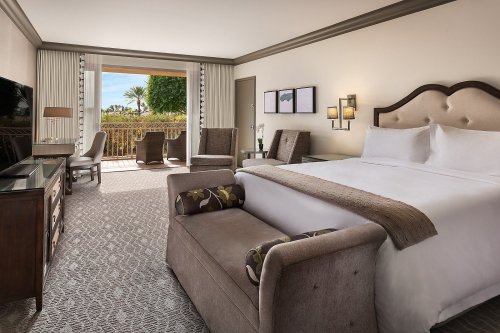 A spacious hotel room featuring a king bed, seating area, and a balcony with views of palm trees and blue skies.